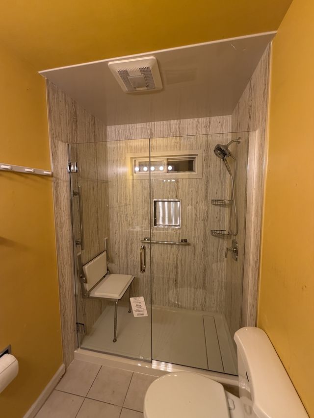 Remodeled walk-in shower with a glass door, stone-pattern walls, a corner bench, and a grab bar.