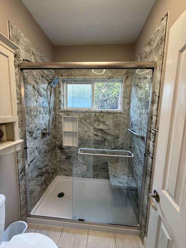 Spacious walk-in shower with stone-pattern walls, a glass enclosure, a corner window, and a grab bar.