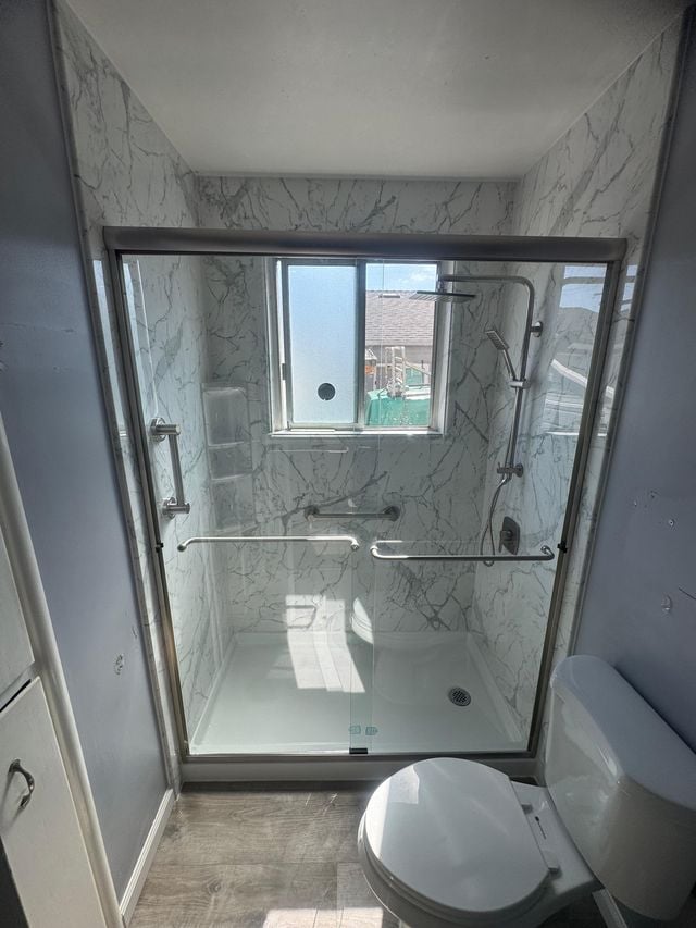 Walk-in shower with sliding glass doors, marble-pattern walls, and a corner window beside a toilet.