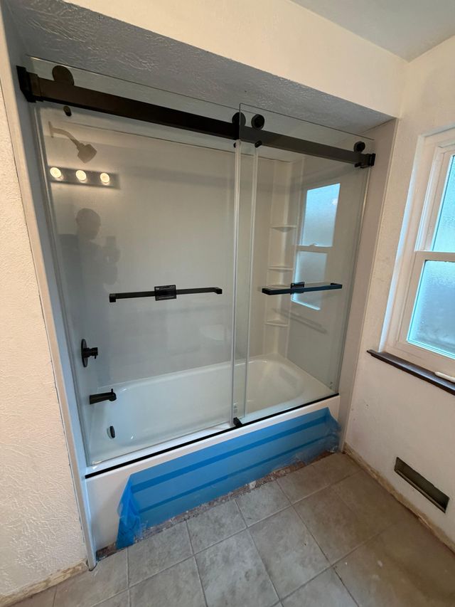 Bathtub fitted with new black-framed sliding glass shower doors and blue floor protection during installation.