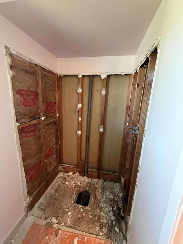 Shower area during demolition with exposed framing, backer board, and an open drain.